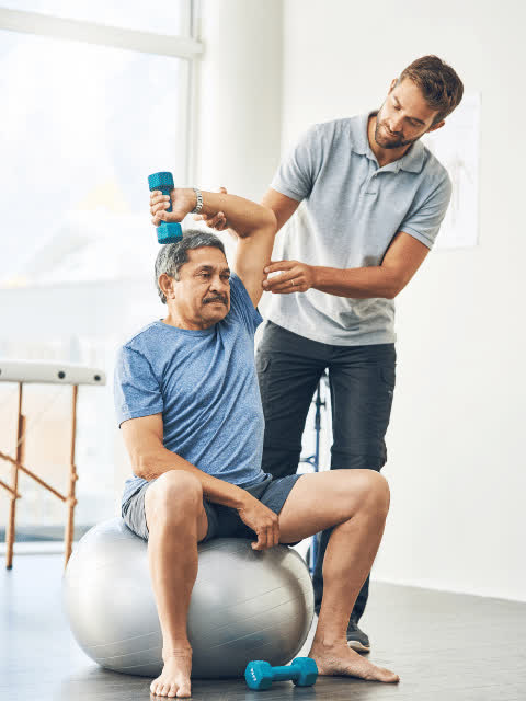 Physiotherapist guiding a patient who lifts a dumbbell while seated on a stability ball